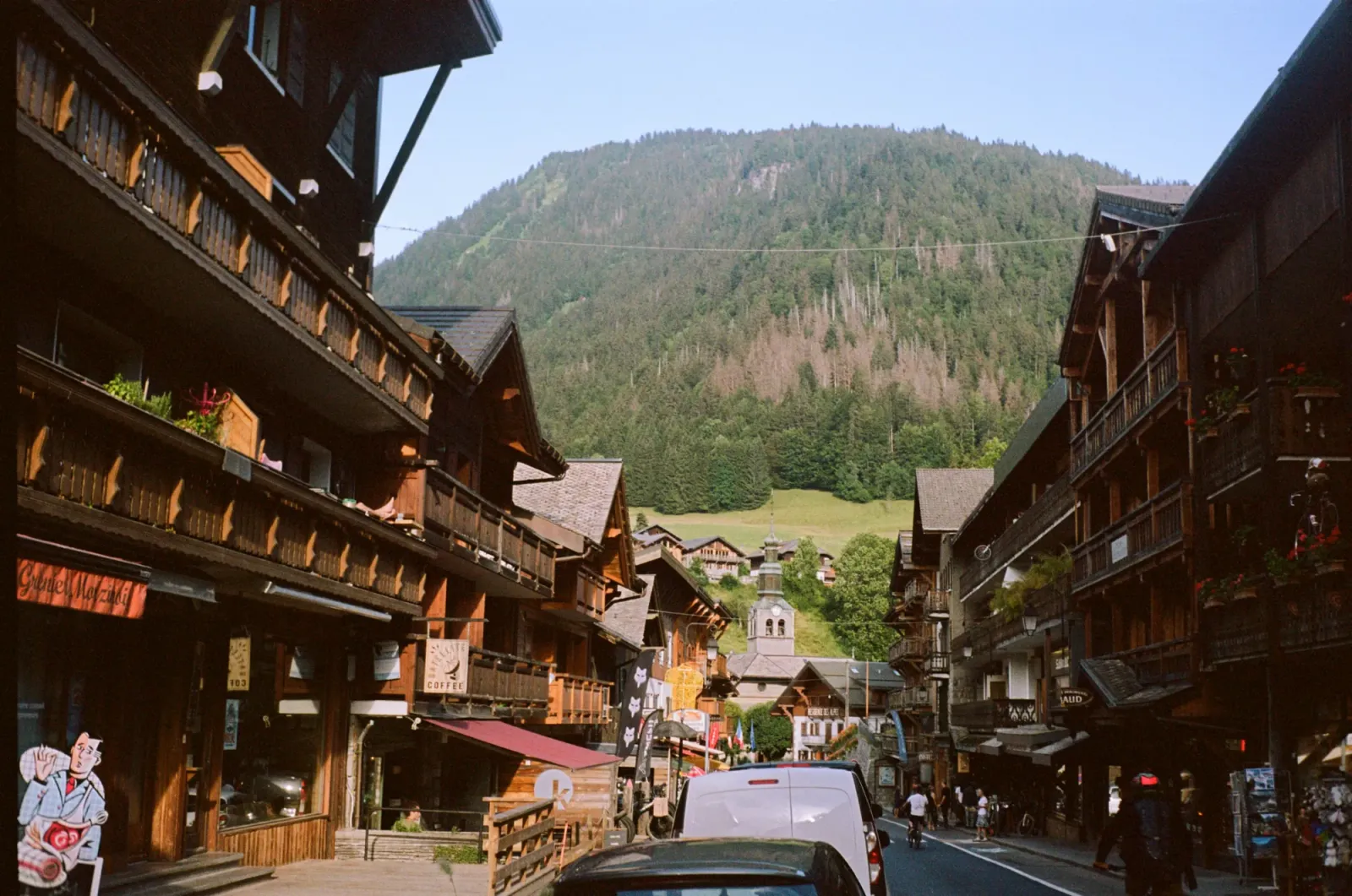 Chalet in legno lungo la via principale di Morzine d’estate, con la chiesa e una montagna boscosa sullo sfondo